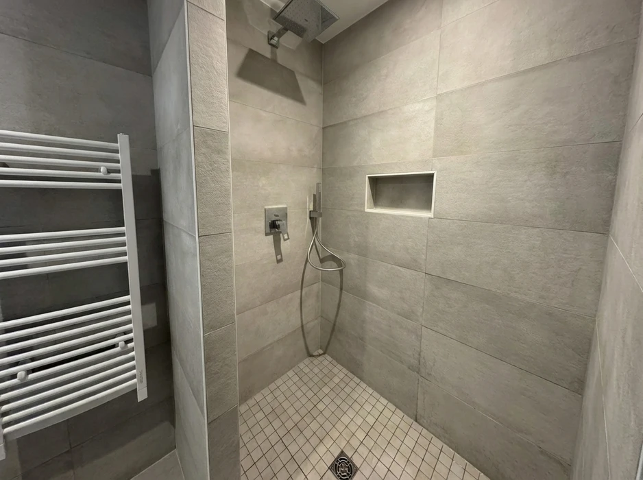 Douche à l'italienne sans porte, carrelage grand format gris béton ciré en pose décalée, niche encastrée dans le mur, mitigeur thermostatique encastré chromé avec douchette flexible, receveur en mosaïque blanche, pommeau de douche pluie au plafond